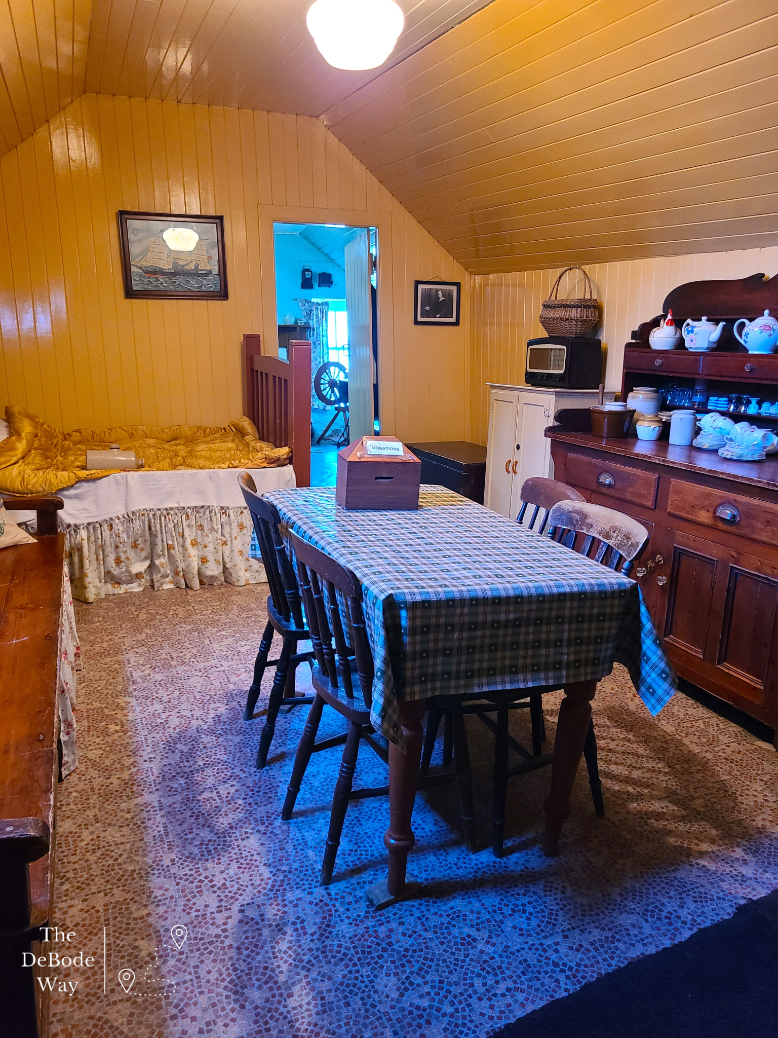 Yellow painted paneling, kitchen table, sideboard with tea fixings, a cupboard and bed in a blackhouse