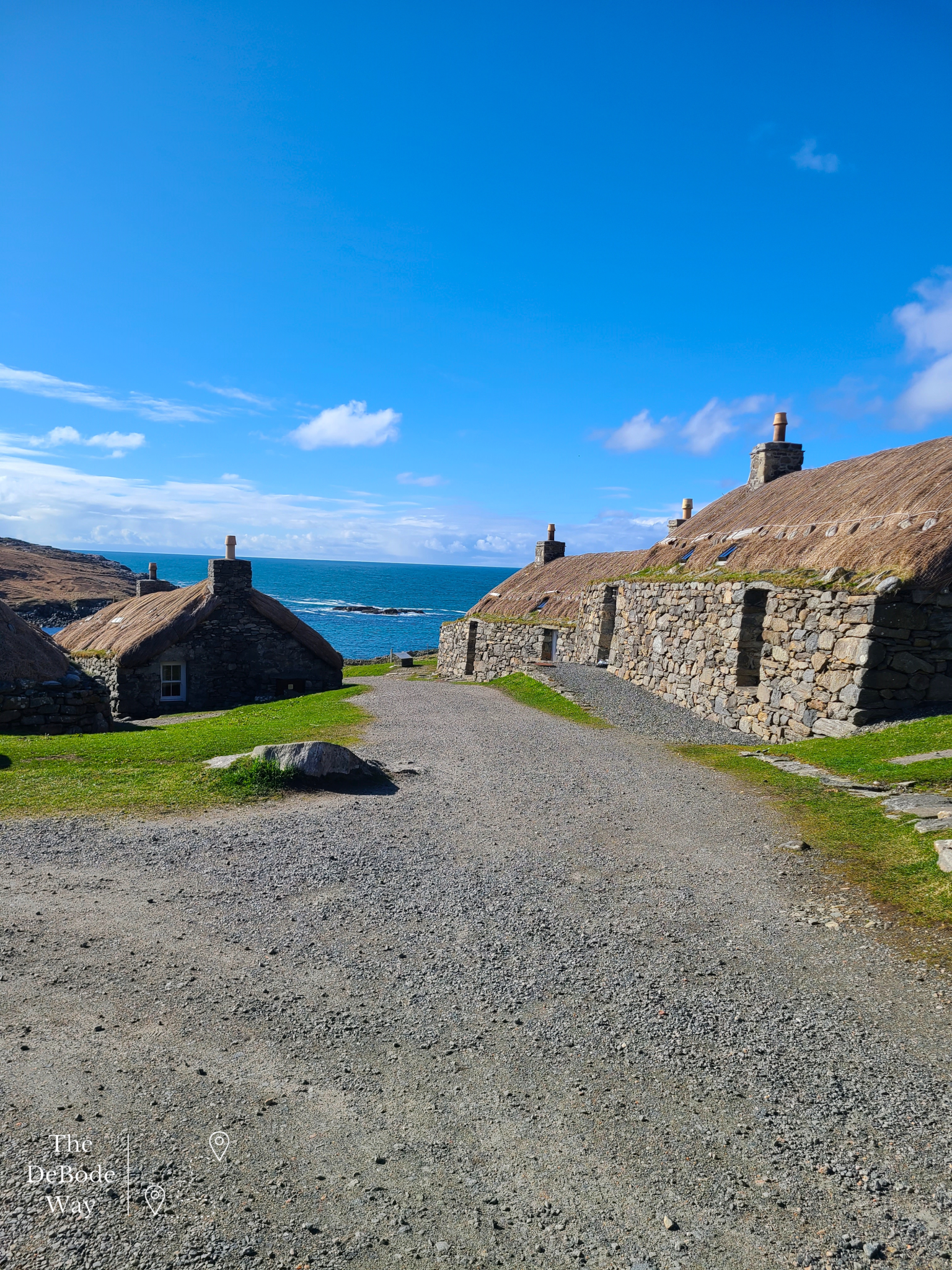 Gravel street lined by stone blackhouses with thatched roofs against a blue sky with the sea in the background