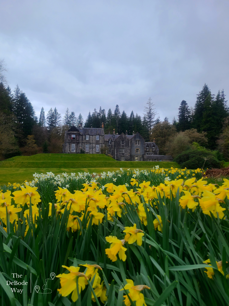 Stone clad Ardanaiseig Hotel under a cloudy sky situated on a green mound with daffodils below.