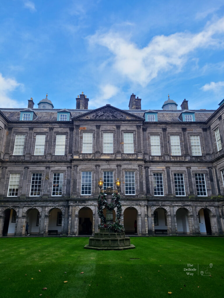 Inner courtyard of Holyrood Palace with green grass below, three stories of stone building and blue sky above.
