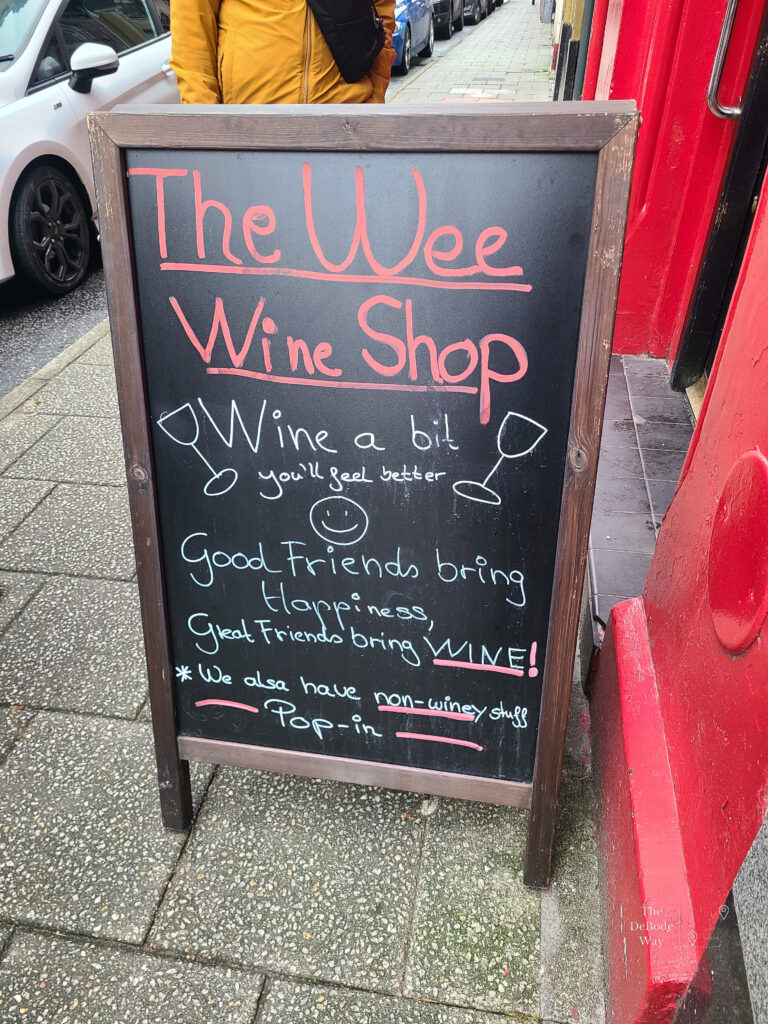 Chalkboard street sign for the Wee Wine Shop that says "wine a bit, You'll feel better" "good friends bring happiness, great friends bring wine" "we also have no-winey stuff, pop in"