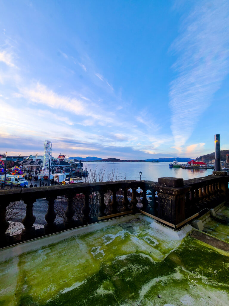 The view from the 1st floor of the Perle Oban Hotel across the bay - mountains across the bay with stone railing green with algae in the foreground. CalMac ferry pier to the left and fishing boats to the right. Oban, Scotland