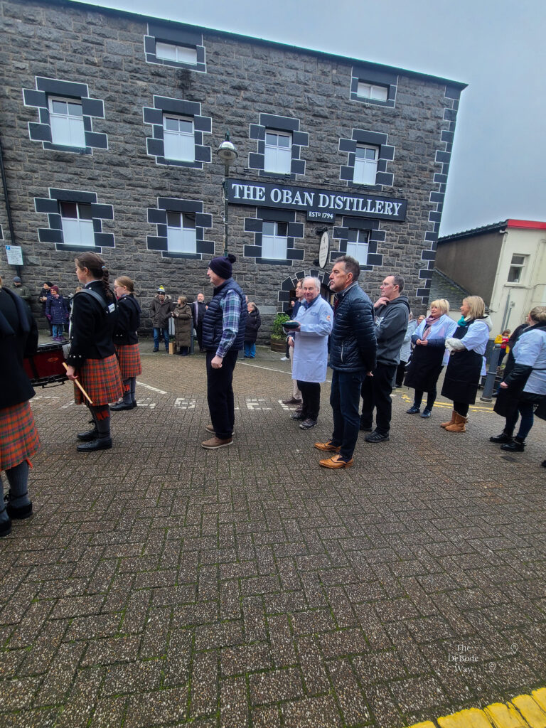 Oban high school pipe band leading in the golden haggis winner from the previous year followed by chefs in white coats who are contenders for this year's competition. In front of Oban Distillery.