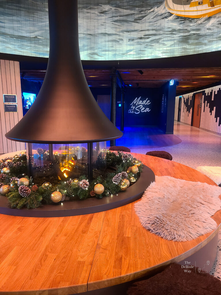 Circular fireplace with wood seating surround decorated with garland, pine cones and gold baubles with "Made by the Sea" on the wall behind
