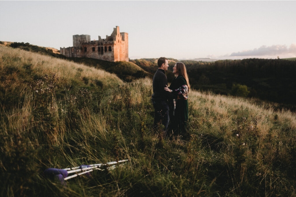 Jason and Tasha looking at each other on a grassy hillside with a castle ruin in the background. Crutches in the grass in the foreground.